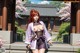 A woman in traditional attire stands on steps with a sword, cherry blossoms and a temple in the background.